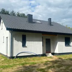 Nowy dom szkieletowy na lata, z szarym dachem i białymi ścianami, otoczony zielenią i lasem.