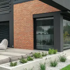 Nowoczesny projekt domu z dużymi oknami, ceglaną elewacją i minimalistycznym ogrodem. Wygodny szezlong zaprasza do relaksu.