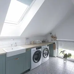 Jasna pralnia na poddaszu z pralką, suszarką, dużą umywalką i oknem dachowym.