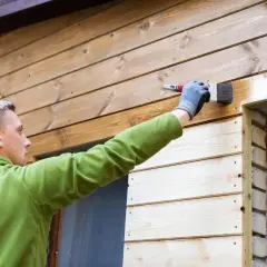 Mężczyzna w zielonej bluzie maluje pędzlem drewnianą elewację domu, nadając jej nowy wygląd.