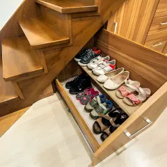 Zabudowa pod schodami z wysuwanymi szufladami na buty. Widać różne rodzaje obuwia, od dziecięcych sandałków po sportowe buty.
