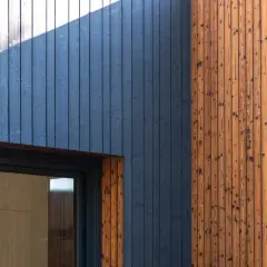 Nowoczesna elewacja z ciemnoniebieskich desek i naturalnego drewna. Widoczny fragment okna i ruszt pod elewację drewnianą.