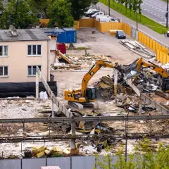Dwa koparki pracują na placu budowy, rozbierając budynek. W tle widać drogę z samochodami i drzewa.