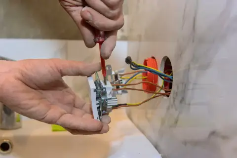 Ręce montują gniazdko, przykręcając je śrubokrętem. Widać kolorowe przewody wychodzące z puszki instalacyjnej.