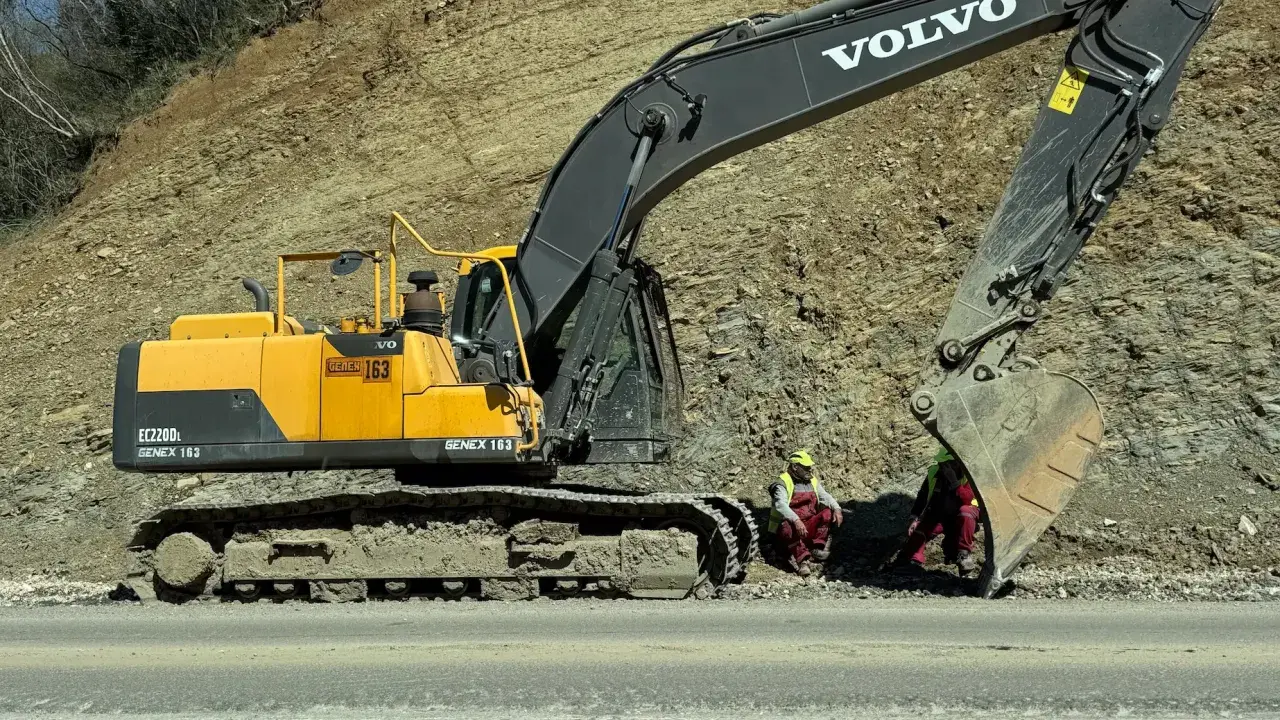 Dwa rodzaje koparek, w tym żółta Volvo EC220DL, pracują przy osuwisku. Dwóch robotników w kamizelkach odblaskowych odpoczywa.