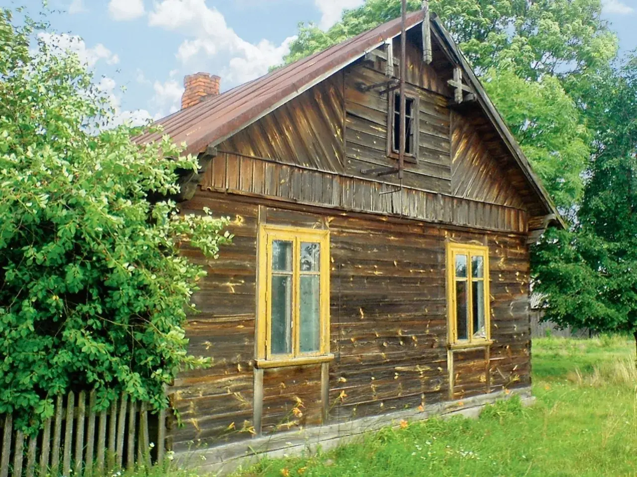 Stary, drewniany dom z żółtymi oknami, otoczony zielenią. Wnętrze tego domu kryje zapewne wiele historii.
