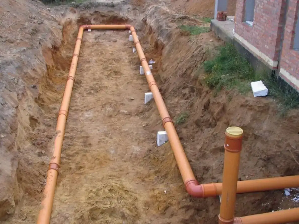 Instalacja gruntowego wymiennika ciepła w wykopie. Pomarańczowe rury układane są w ziemi obok budowanego domu.