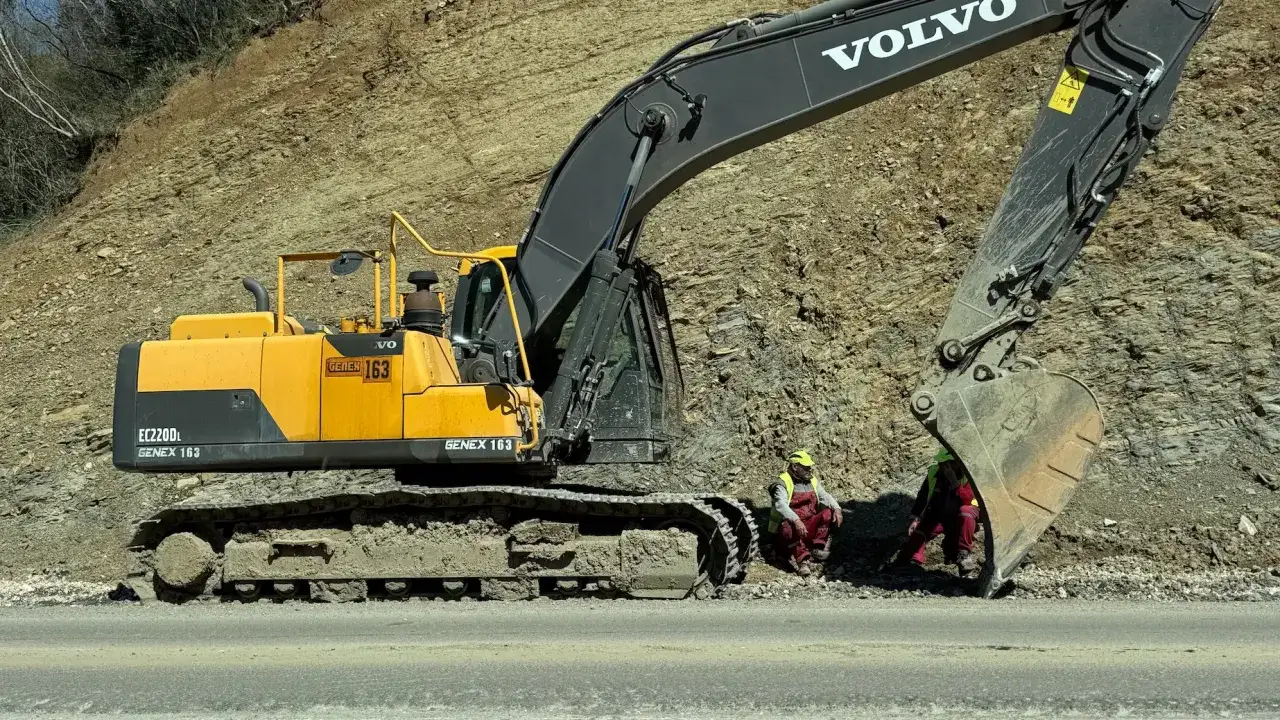 Żółta koparka Volvo EC220DL, jeden z wielu rodzajów koparek, pracuje przy osuwisku. Dwóch robotników w kamizelkach odblaskowych odpoczywa obok.