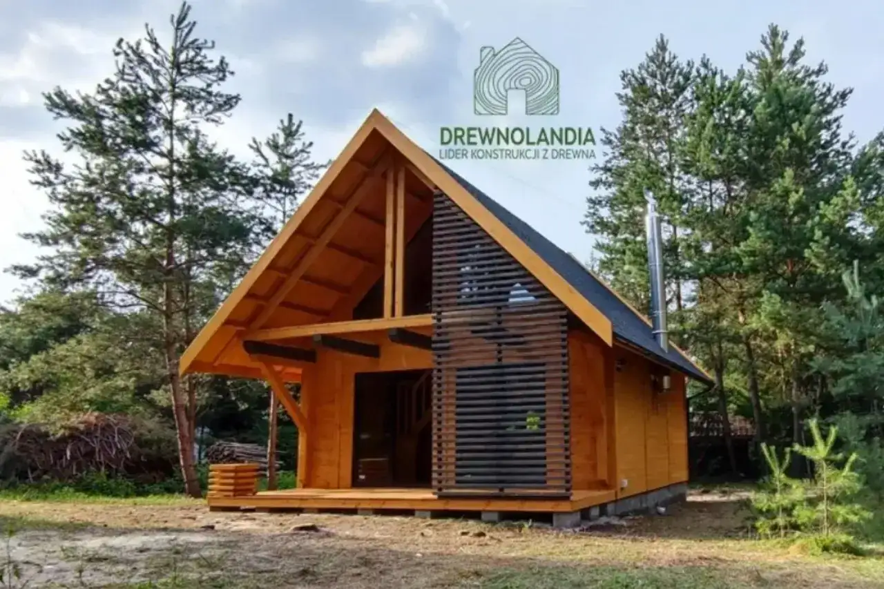 Drewniany domek letniskowy w lesie, z charakterystycznym dachem i drewnianą elewacją. Trwa budowa, widać elementy konstrukcyjne.