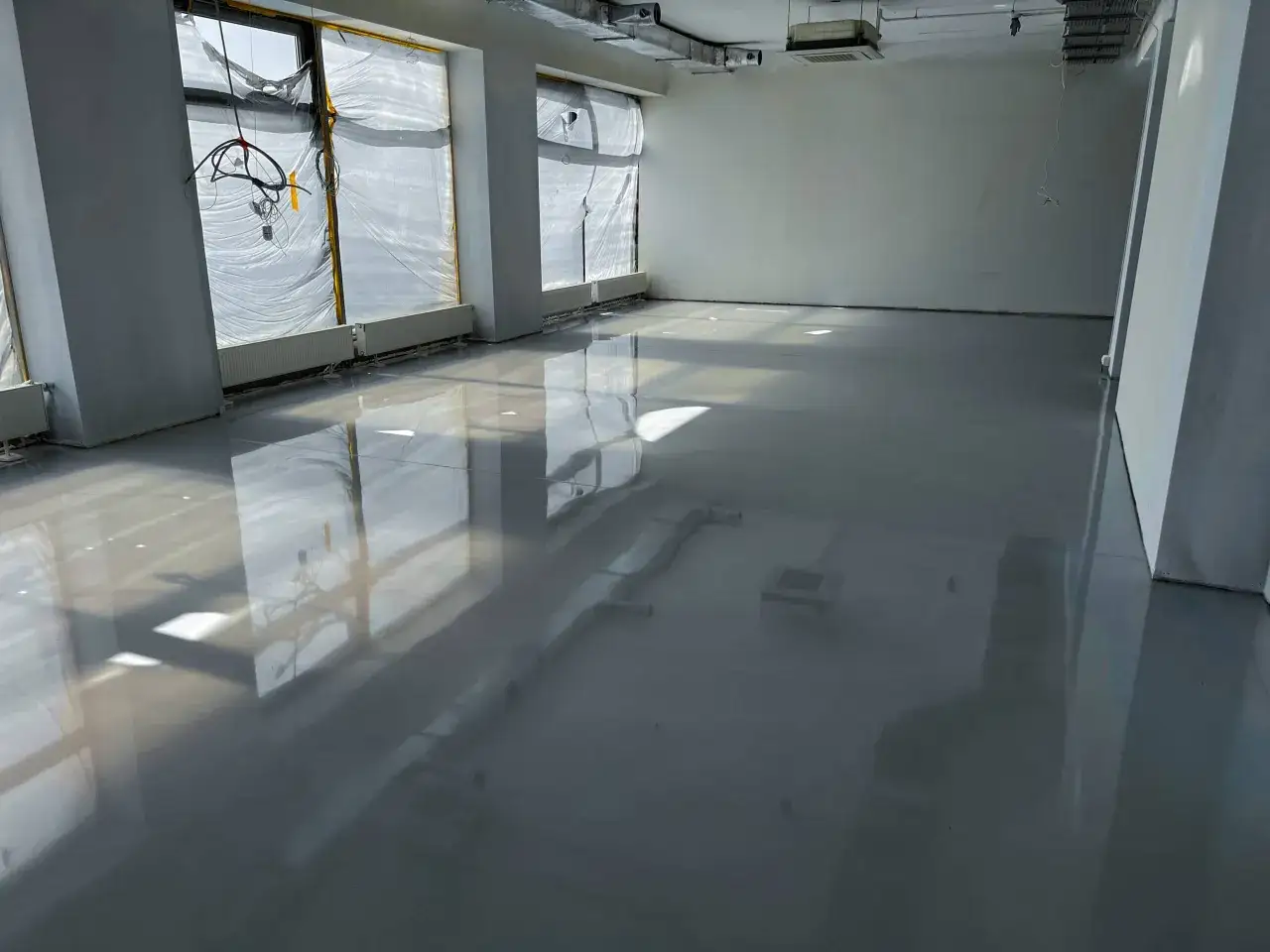 Jasna, lustrzana podłoga epoksydowa odbija światło i okna z folią ochronną. Widać instalacje wentylacyjne i grzejniki.