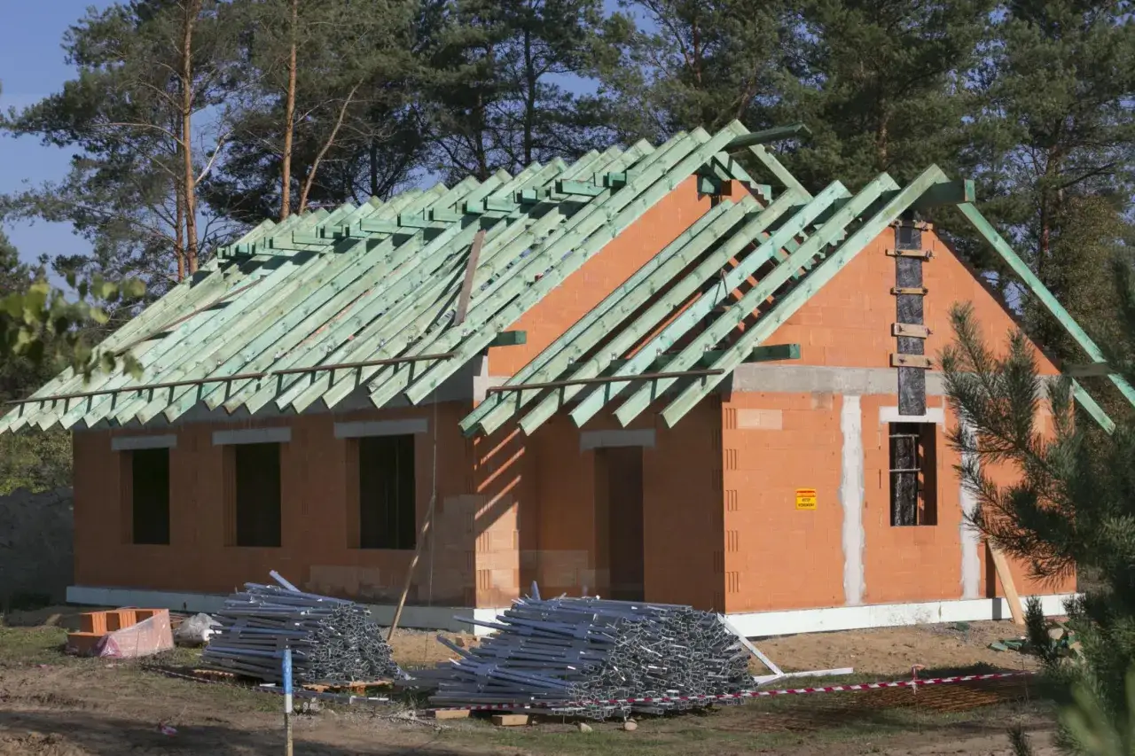 Dom w budowie, widać ceglane ściany i zieloną więźbę dachową. Materiały budowlane leżą na ziemi. To etap samodzielnej budowy domu.