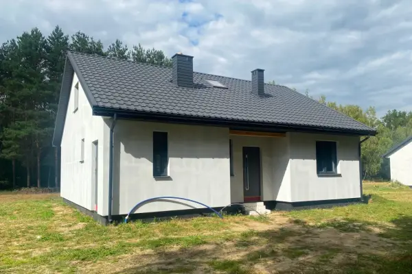 Nowy dom szkieletowy na lata, z szarym dachem i białymi ścianami, otoczony zielenią i lasem.
